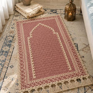 Op de afbeelding: Een rechthoekig gehaakt gebedskleed in oudroze en crème met een centraal mihrab-boogontwerp. De rand bevat een crèmekleurig ruitpatroon en een rij crèmekleurige kwastjes langs de onderkant, vervaardigd met getextureerd garen.