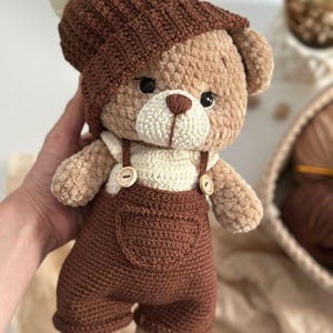 May include: A handmade crochet teddy bear wearing a brown hat with a cream pom-pom and matching brown dungarees. The bear has a cream shirt and wooden button details. The bear's fur is a light brown colour.