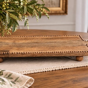 May include: A rectangular wooden serving board with a natural brown finish, featuring a beaded edge detail, a short handle on one side, and four small spherical feet that elevate the board slightly above the surface it is placed upon.