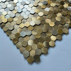 Puede incluir: Primer plano de una sección de baldosas hexagonales metálicas. Las baldosas son en tonos dorados y plateados, creando un patrón geométrico texturizado. Las baldosas están dispuestas en un diseño de panal, adecuado para aplicaciones decorativas.