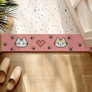 May include: A pink rectangular doormat featuring pixelated cat faces, paw prints, and a heart design. The mat is placed on a wooden floor, with a pair of beige slippers nearby. The doormat is a decorative item.