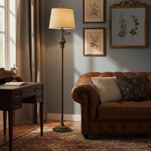 May include: A bronze floor lamp with a beige lampshade stands in a living room. The lamp has a decorative base and a curved arm. A brown leather sofa with pillows is in the background, along with framed botanical prints and a wooden desk.