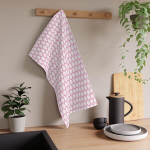 Puede incluir: Un paño de cocina con estampado rosa y blanco cuelga de un perchero de madera. El paño presenta un diseño repetitivo de formas blancas sobre un fondo rosa. El paño de cocina está hecho de un material suave y absorbente, ideal para secar platos o limpiar superficies.