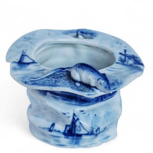 May include: A blue and white ceramic decorative container with a nautical theme. The container features a stylized seal figure and painted scenes of sailboats and windmills. The rim has a unique, curved shape.