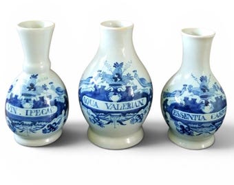 May include: Three antique white ceramic apothecary bottles with blue floral and text designs. The bottles have a bulbous shape with a narrow neck and a flared rim. The text on the bottles reads "VIN. IPECAC", "AQUA VALERIAN", and "ESSENTIA CASTO".