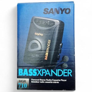 Può includere: Un lettore radio/cassette stereo personale Sanyo BassXpander vintage nella sua scatola originale. La scatola è blu con il logo Sanyo e i dettagli del prodotto. Il lettore è nero con un quadrante argentato e il logo BassXpander.