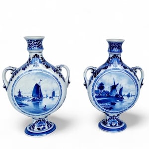 Pode incluir: Dois vasos de cerâmica azuis e brancos a condizer. Cada vaso apresenta um painel central redondo que retrata uma cena náutica com veleiros e moinhos de vento. Os vasos têm uma forma bulbosa com alças e padrões florais.