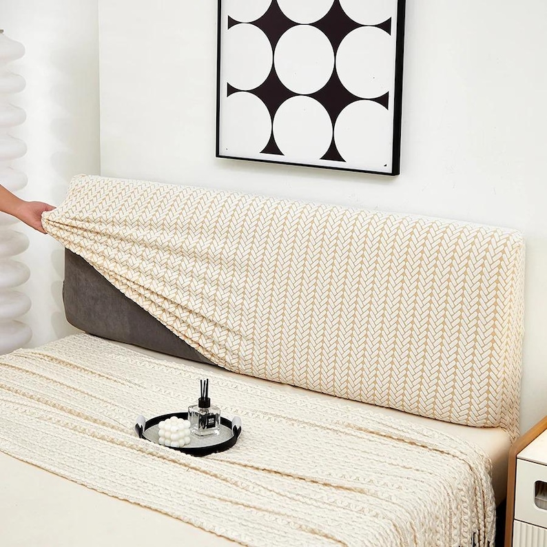 May include: A beige headboard cover with a repeating geometric pattern is being placed on a gray headboard. A matching blanket and decorative tray with a candle and perfume bottle are on the bed. A black and white abstract art piece hangs on the wall.