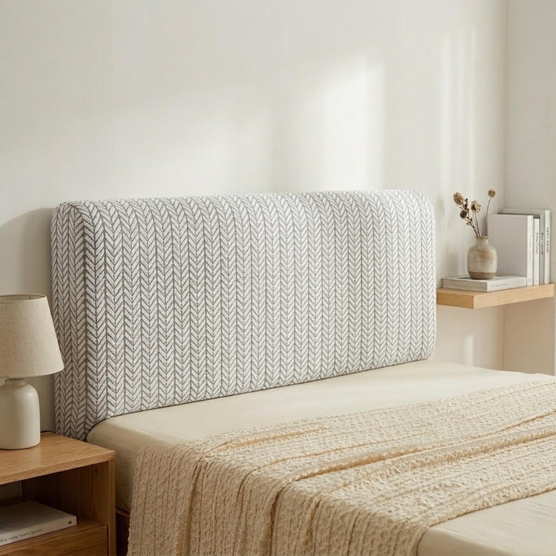 May include: A rectangular bed headboard featuring a grey and white chevron design. The headboard is padded, with a neutral-toned bedspread. A wooden bedside table with a lamp is also visible.