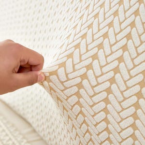 May include: Close-up of a beige and white patterned fabric. The fabric has a textured, woven design with a repeating geometric pattern. A hand is pulling the fabric, showing its stretch and texture. The background is a lighter, similar fabric.