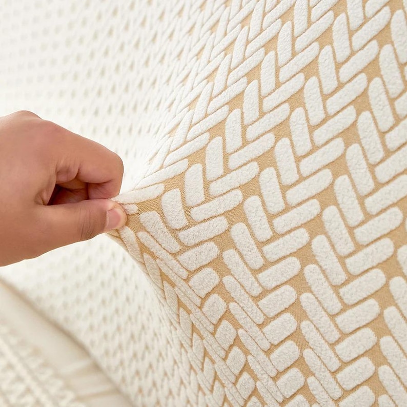 May include: Close-up of a beige and white patterned fabric. The fabric has a textured, woven design with a repeating geometric pattern. A hand is pulling the fabric, showing its stretch and texture. The background is a lighter, similar fabric.
