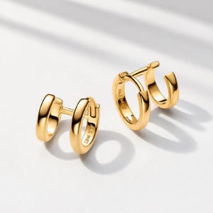 May include: A pair of gold-toned hoop earrings featuring a double-hoop design. Each earring consists of two connected circular bands with a polished, smooth finish. The metal surface reflects light, creating highlights along the curved edges.