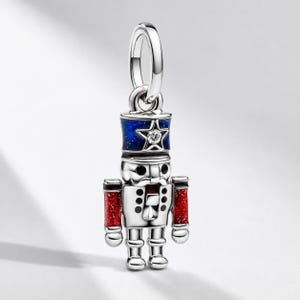 May include: A silver charm shaped like a nutcracker soldier featuring a blue hat with a white star and a clear stone, red glittery sleeves, and black painted details on the face and torso, suspended from a circular silver jump ring bail.
