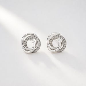 May include: A pair of silver-coloured stud earrings featuring an interlocking circular design. One circle is smooth and polished, while the other is encrusted with small, clear, sparkling stones arranged in a continuous pattern around the ring.