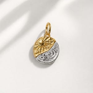 Puede incluir: Un colgante circular bicolor que presenta un motivo de sol dorado en la mitad superior y un diseño de luna creciente y estrellas plateadas en la mitad inferior. El colgante incluye una pequeña anilla circular en la parte superior.
