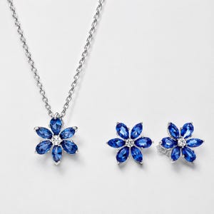 Può includere: Un set di gioielli color argento composto da una collana con pendente e due orecchini a lobo. Ogni pezzo presenta un design a forma di fiore con sei pietre blu taglio marquise che circondano una pietra rotonda trasparente al centro.