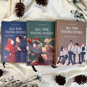May include: Three hardcover books titled All The Young Dudes by mskingbean89. The blue book shows a couple dancing, the green book depicts two people lying down, and the brown book features three figures sitting together on a stone ledge.
