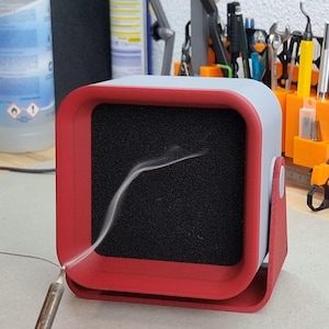 Könnte beinhalten: Ein rot-grauer Lötrauchabsauger mit einem schwarzen Filter. Das Gerät ist quadratisch mit abgerundeten Ecken und einem roten Sockel. Rauch ist sichtbar, was auf die Verwendung hindeutet. Werkzeuge und Zubehör sind im Hintergrund.