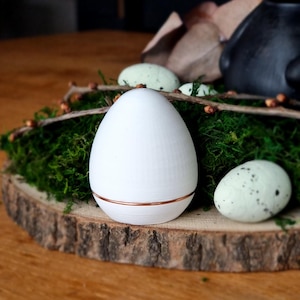 Könnte beinhalten: Ein weißes Ei mit kupferfarbenem Band liegt auf einer Holzscheibe mit grünem Moos, Zweigen und gesprenkelten Eiern. Eine rustikale Oster- oder Frühlingsdekoration.