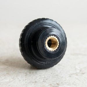 May include: A black, round knob with a ridged, textured surface. The center features a brass-colored threaded insert. The knob is likely for furniture or a similar application.