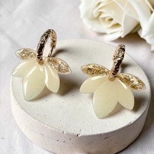 Peut inclure: Une paire de boucles d'oreilles créoles avec un anneau en métal doré texturé attaché à une breloque en résine jaune pâle en forme de pétale. Le haut de chaque breloque est orné de flocons métalliques dorés, créant un effet scintillant.