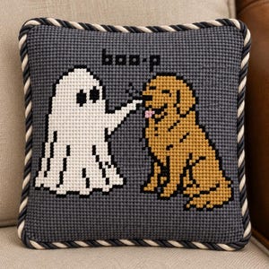 May include: A square decorative pillow featuring a dark gray needlepoint background with a white ghost touching the nose of a golden-colored dog. The text "boo-p" is stitched in black above the figures. The pillow has a twisted black and white cord trim.