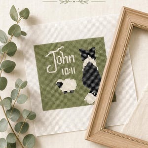 May include: A cross-stitch embroidery on white fabric featuring a green background with a black and white dog, a small white sheep, and the text "John 10:11" in white. The piece is displayed alongside a wooden frame and a green eucalyptus branch.