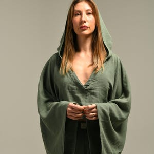 Può includere: Un top verde con cappuccio con maniche larghe e scollo a V profondo. Il top è abbinato a pantaloni abbinati. L'outfit è realizzato in un tessuto morbido e fluido. La modella indossa un anello.