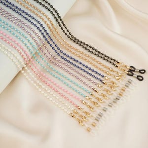 Peut inclure: Plusieurs chaînes à lunettes perlées de différentes couleurs, dont blanc, rose, bleu et noir. Chaque chaîne est ornée de petites perles rondes et de fermoirs métalliques pour attacher les lunettes. Les chaînes sont présentées sur un tissu blanc et soyeux.