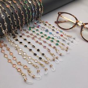 Peut inclure: Plusieurs chaînes à lunettes avec des pierres précieuses rondes et colorées serties dans du métal doré. Les chaînes sont présentées avec une paire de lunettes écailles. Les chaînes ont des boucles en caoutchouc blanc pour les attacher aux lunettes.