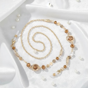 Op de afbeelding: Goudkleurige brillenketting met bruine en heldere kralen, en kleine goudkleurige cirkelvormige accenten. De ketting wordt gepresenteerd op een witte, zijdeachtige ondergrond, met verspreide imitatieparels.
