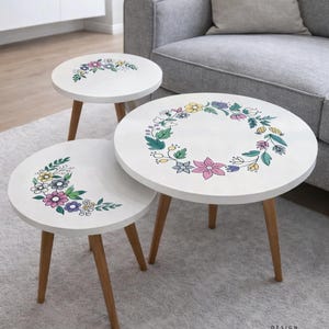 May include: Set of three white round side tables with wooden legs. Each table features a floral design in pastel colors. The tables are arranged in a nested configuration, offering a stylish and functional accent for any living space.