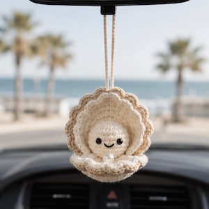 May include: Crocheted car charm featuring a smiling pearl inside a beige seashell. The charm is suspended from a rearview mirror. The background shows a beach scene with palm trees and the ocean.