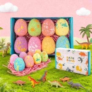 Puede incluir: Un juego de regalo de bombas de baño con temática de dinosaurios en forma de huevo, presentado en una caja azul. El juego incluye varios colores y texturas, con pequeñas figuras de dinosaurios esparcidas. También hay una caja con las palabras "BATH BOMB".