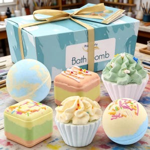 Könnte beinhalten: Eine Geschenkbox mit hellblauem Aquarelldesign und goldenem Band, die mehrere Badekugeln enthält. Die Badekugeln haben verschiedene Formen, darunter Cupcakes und Kugeln, und sind mit farbenfrohem Zuckerguss und Streuseln verziert.