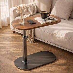 May include: A brown, oval-shaped side table with a dark metal base and adjustable height. The tabletop holds a coffee cup, book, glasses, a smartphone, and a small vase with dried flowers. The table is positioned next to a beige sofa.