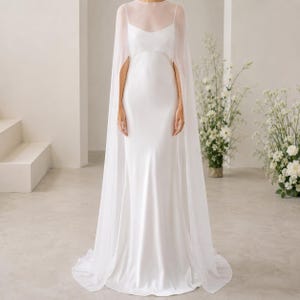 May include: A white, floor-length wedding dress with a fitted bodice and a flowing skirt. The dress is paired with a sheer, cape-like overlay that drapes over the shoulders and arms. The dress is in a bright, well-lit room.