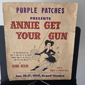 May include: A vintage paper poster for the 1959 production of Annie Get Your Gun at the Grand Theatre. The poster features purple and red text on a tan background, including the title, Irving Berlin, and dates, with a black illustration of a performer.