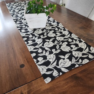 Puede incluir: Un camino de mesa con estampado floral en blanco y negro. El camino de mesa es rectangular y presenta un diseño repetido de flores y hojas estilizadas. Una jardinera de madera blanca con plantas verdes encima.