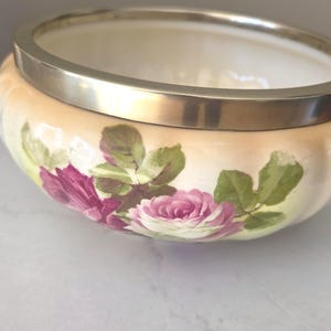 May include: A round porcelain bowl featuring a soft peach and cream gradient background with hand-painted pink rose blossoms and green leaves. The bowl is finished with a smooth, polished silver-toned metal rim around the top edge.