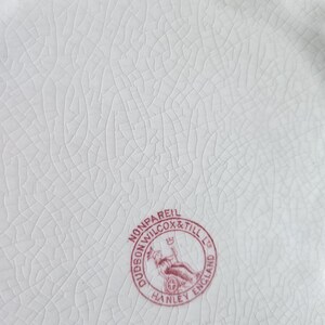 Puede incluir: Primer plano de un plato de cer&aacute;mica blanca con una superficie agrietada y texturizada. Un sello circular rojo dice "Dudson Wilcox & Till Ltd Hanley England Nonpareil". El plato tiene un borde festoneado.