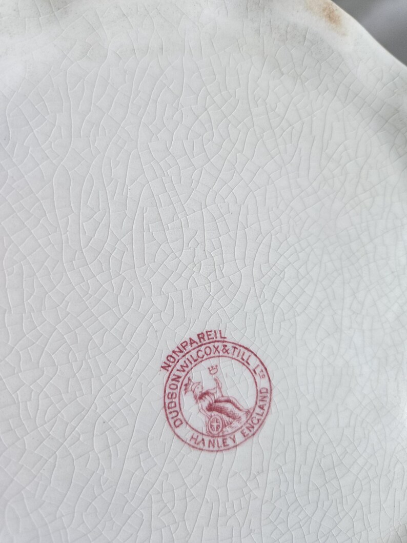 Puede incluir: Primer plano de un plato de cer&aacute;mica blanca con una superficie agrietada y texturizada. Un sello circular rojo dice "Dudson Wilcox & Till Ltd Hanley England Nonpareil". El plato tiene un borde festoneado.