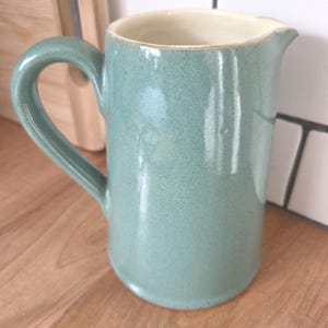 Può includere: Brocca in ceramica con esterno verde acqua chiaro e interno color crema. La brocca ha un manico curvo e un beccuccio. La brocca è appoggiata su una superficie di legno.