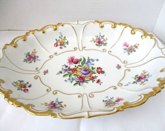 Puede incluir: Un plato ovalado grande de servir blanco con un borde dorado y diseños florales intrincados. El plato está decorado con flores y hojas delicadas y coloridas.
