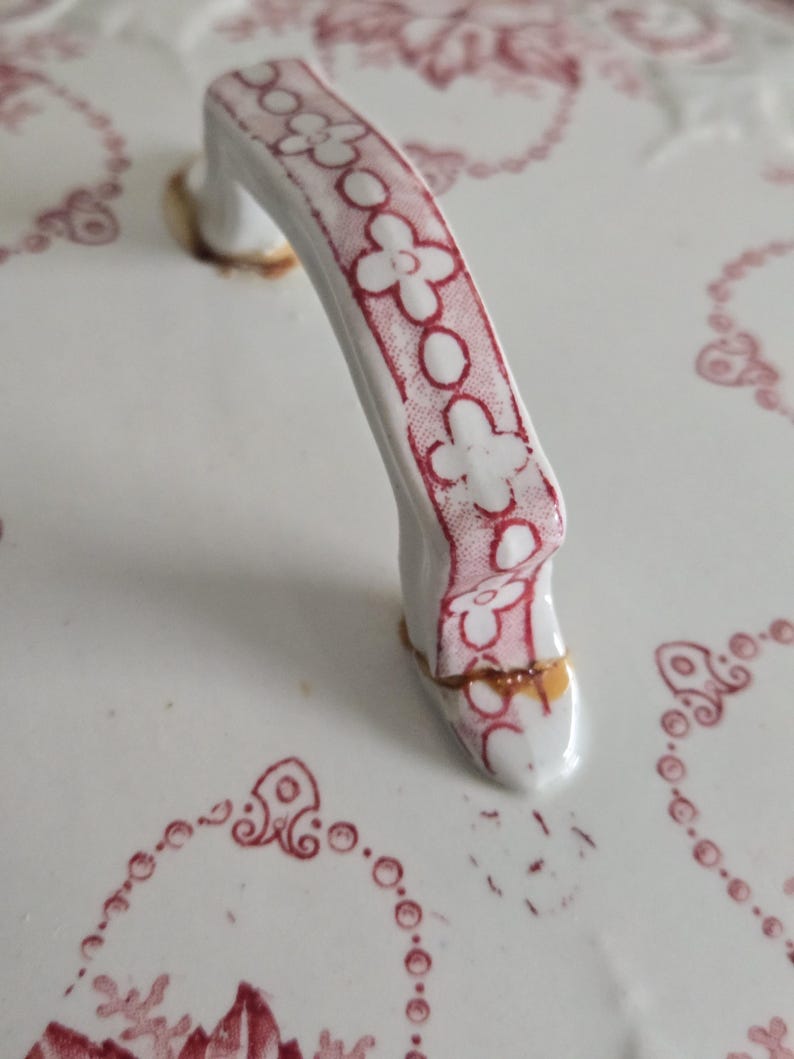 Puede incluir: Primer plano de una tapa de cer&aacute;mica blanca con un asa decorativa. El asa es blanca con un estampado floral rojo. La tapa tiene un dise&ntilde;o floral rojo alrededor de los bordes y es de estilo vintage.