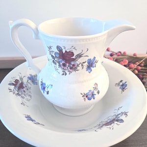 Könnte beinhalten: Ein weißes Keramik-Krug- und Waschbecken-Set. Der Krug hat einen gebogenen Griff und Ausguss, und beide Teile sind mit blauen und violetten Blumenmustern verziert. Das Set ist zum Waschen bestimmt und steht auf einer dunklen Holzoberfläche.