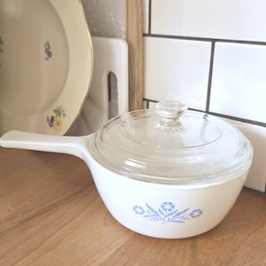 May include: A white ceramic saucepan with a long handle and a clear glass lid. The exterior of the pan features a blue floral pattern consisting of small flowers and leaves centred on the side. The lid has a small, clear glass knob handle.