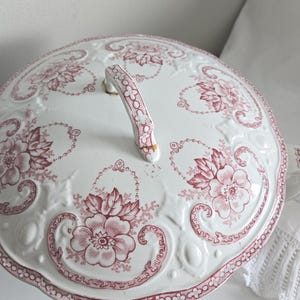 Puede incluir: Tapa de fuente de cer&aacute;mica blanca con un estampado floral rosa. La tapa tiene un asa decorativa y un borde festoneado. El dise&ntilde;o floral incluye flores grandes y peque&ntilde;os detalles florales.
