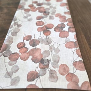 May include: A rectangular table runner with a botanical design. The runner has a cream background with a pattern of eucalyptus leaves in brown and grey. The leaves are arranged in a flowing, organic pattern, suitable for a dining table.
