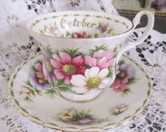 May include: A white teacup and saucer set with a floral design featuring pink, purple, and white flowers. The rim of the cup is inscribed with the word "October" in gold lettering. The set has gold trim.